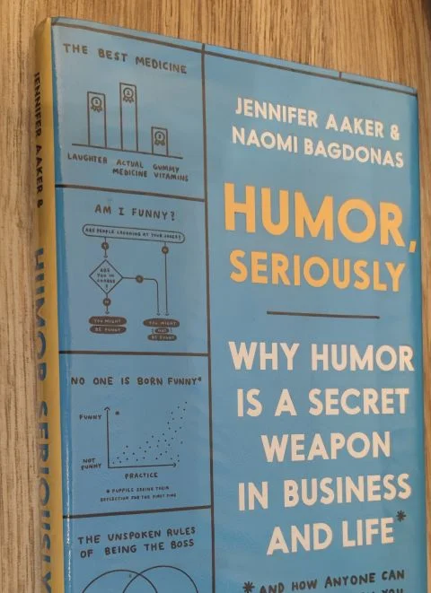 A copy of the book Humor, Seriously by Jennifer Aaker and Naomi Bagdonas, its blue cover covered in hand-drawn cartoon panels, on a wooden table.