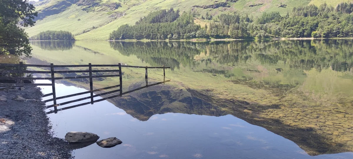 Lake Buttermere on a perfectly still day: a wooden fence runs out into the water, and the hills and trees are mirrored in the lake.